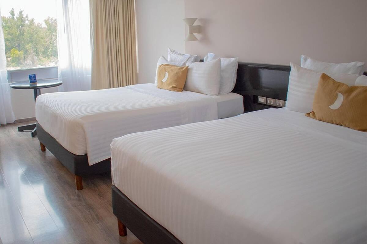 A hotel room with two beds, a small table, and a view of trees through the window.