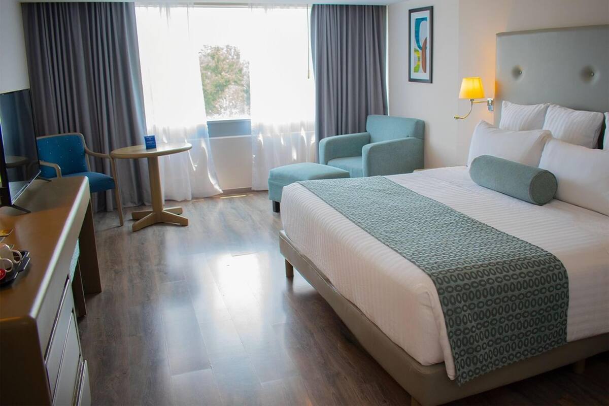 A hotel room with a bed, two armchairs, a small table, a desk, and a window with curtains.
