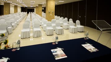 A conference room with rows of white-covered chairs, a long table with water bottles and brochures, and a dark-colored tablecloth.