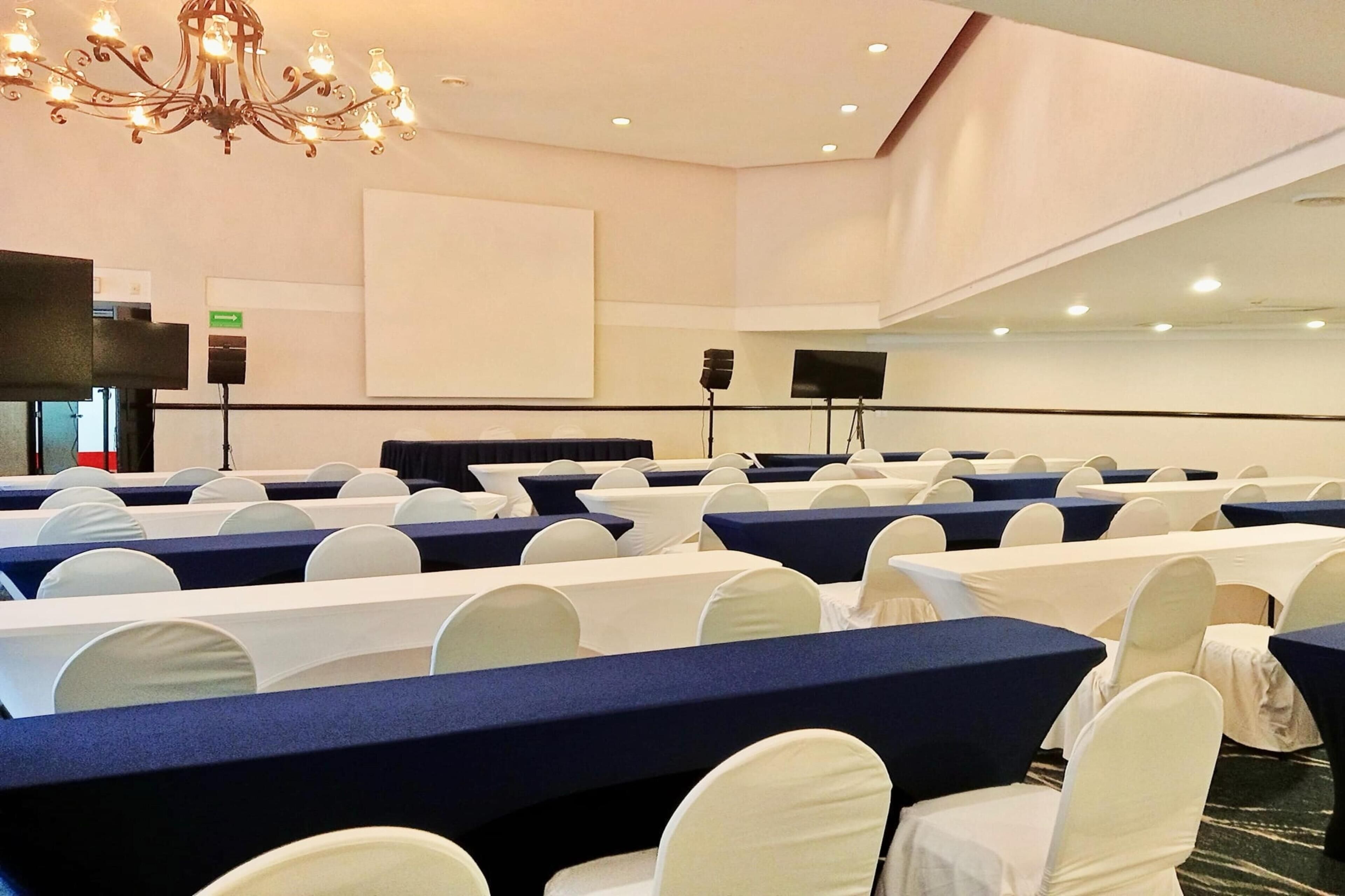A conference room with rows of tables and chairs, a chandelier, and a projection screen.