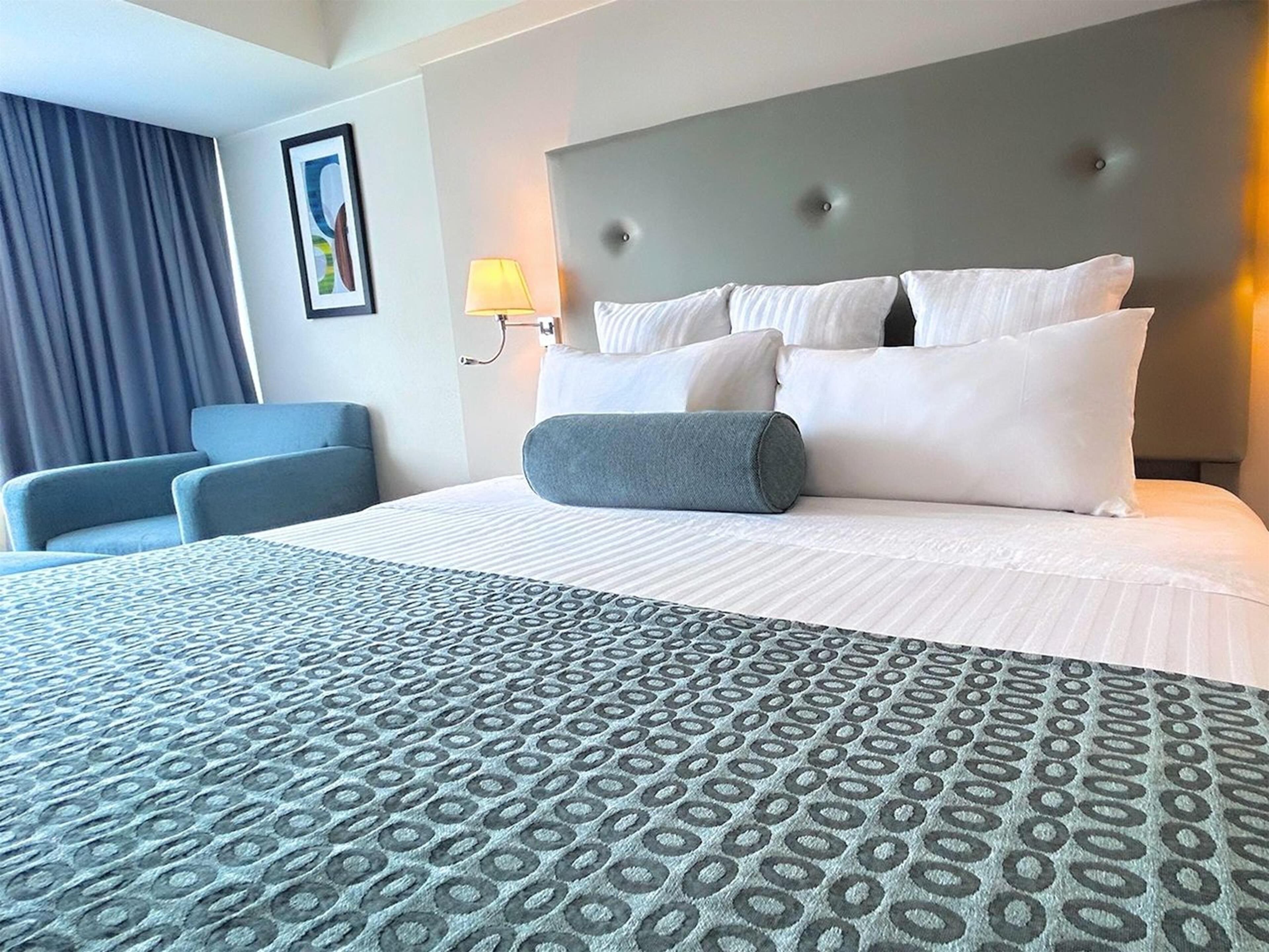 A hotel room with a large bed, a blue armchair, a grey pillow, and a wall-mounted lamp.