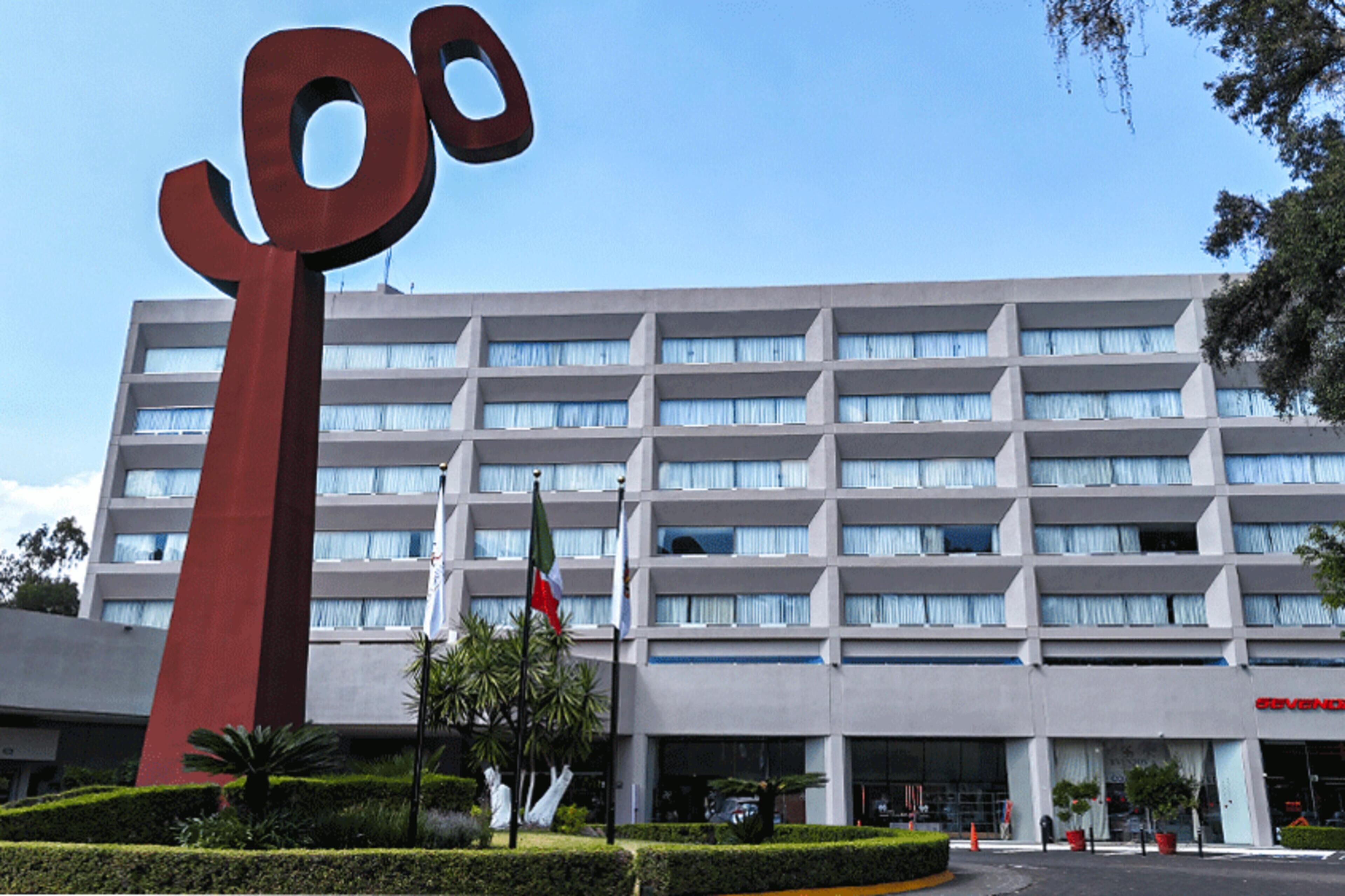 A multi-story hotel with a large red sculpture in front, a flagpole with flags, and a well-maintained garden.