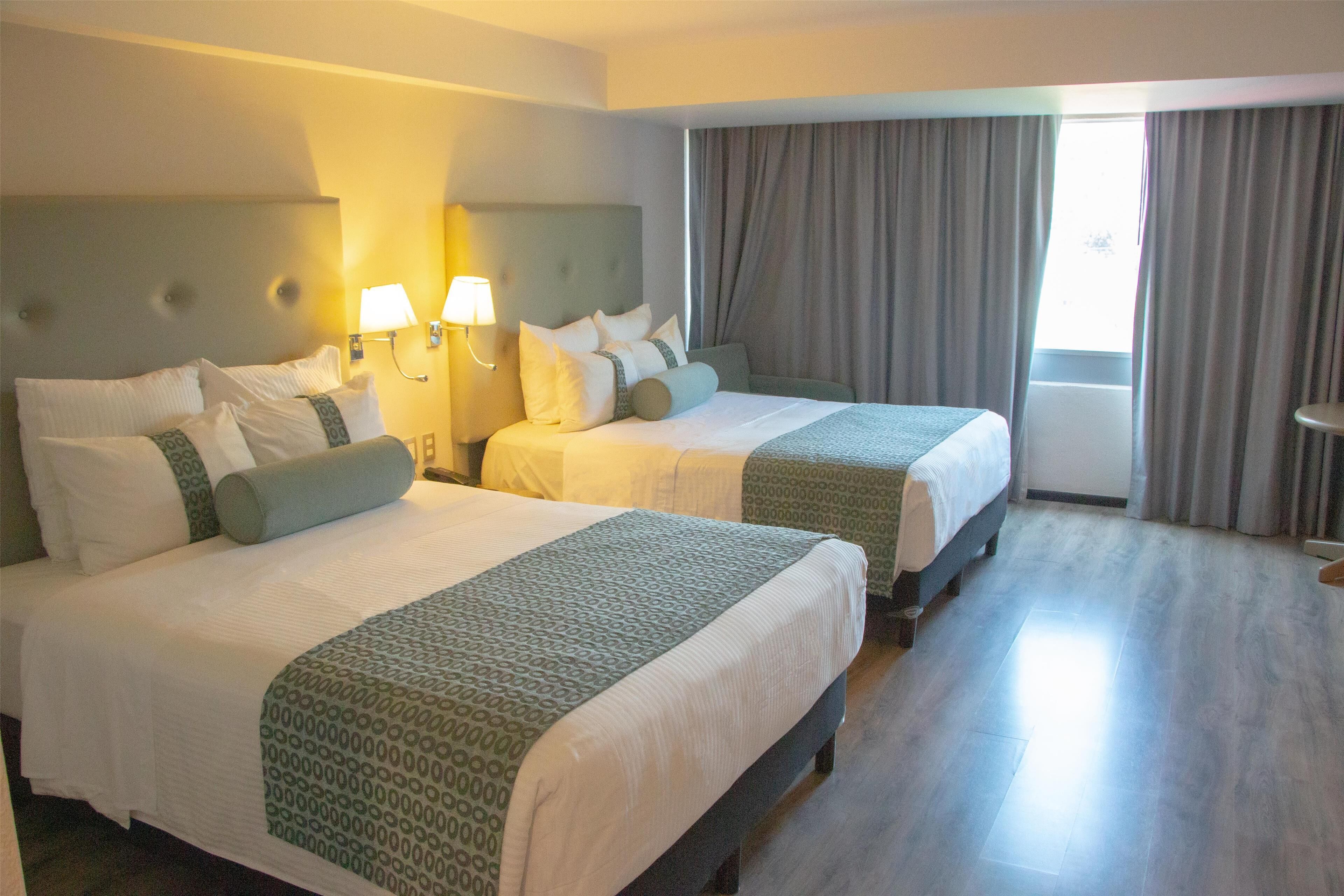 A hotel room with two beds, each with a headboard, bedside lamps, and a window with curtains.