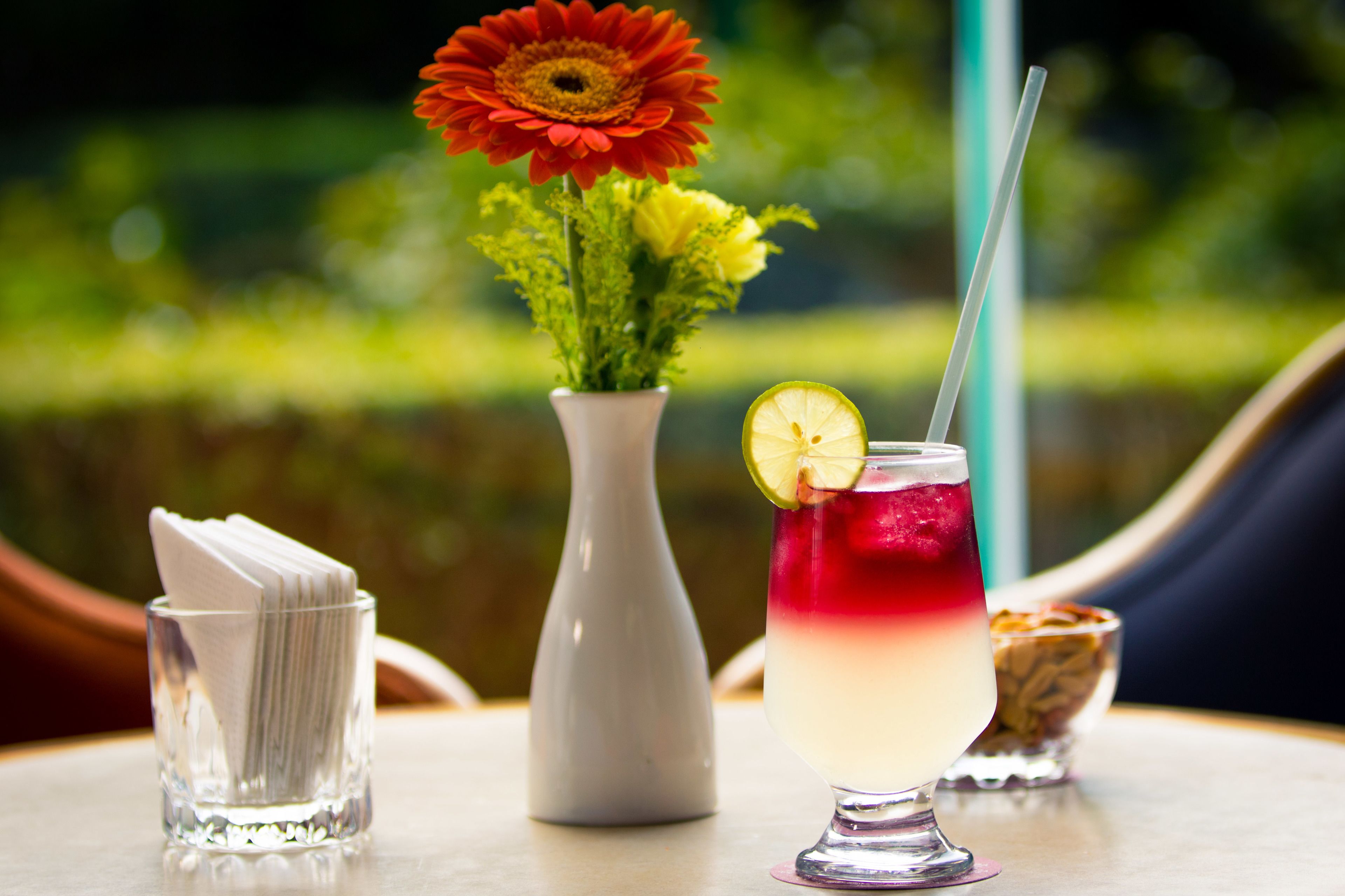 Una mesa con un vaso de bebida en capas, un florero con flores y un reposa servilletas.