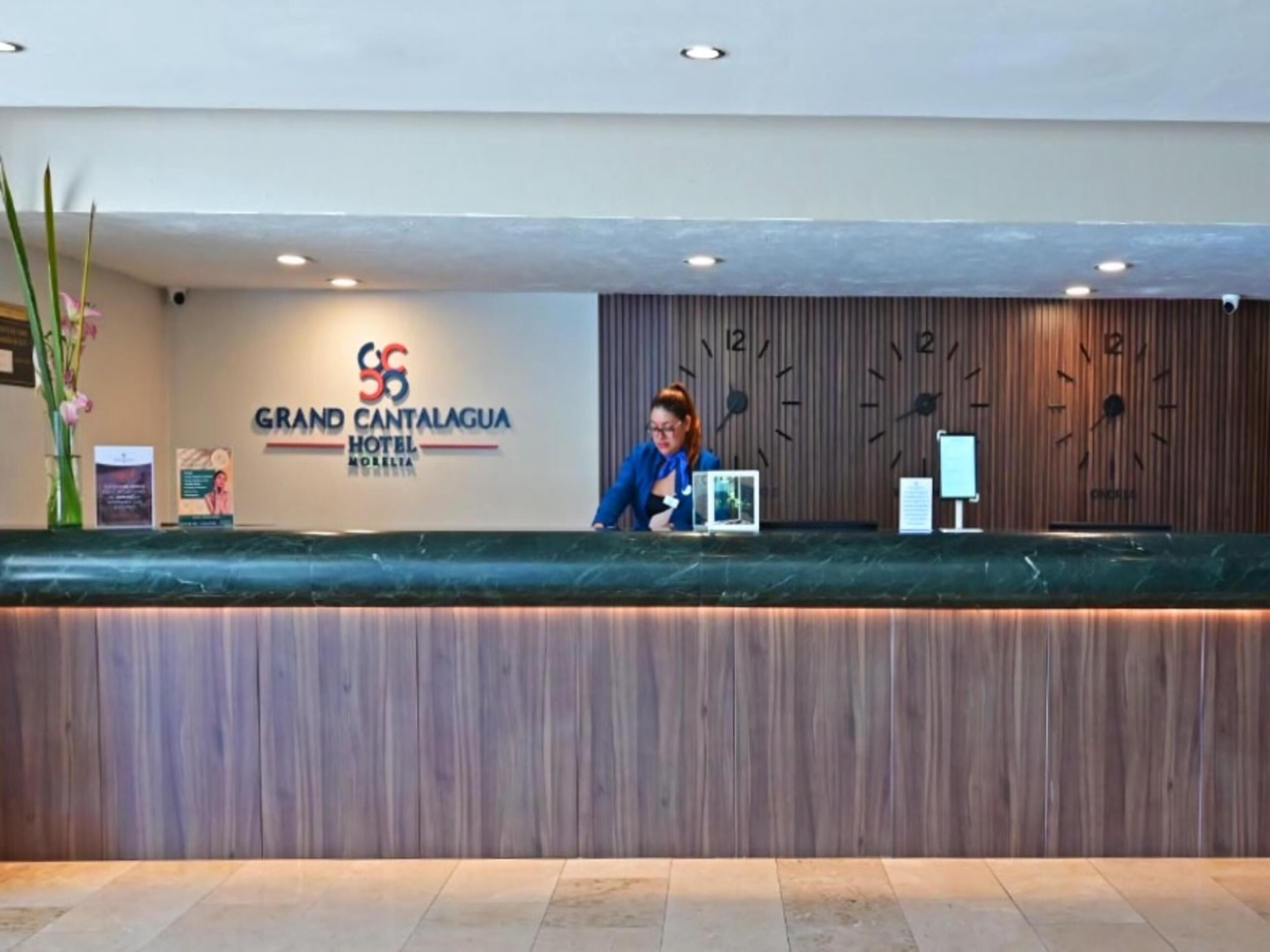 Mostrador de recepción en el Hotel Grand Cantagua, con un panel de pared de madera, un reloj y una pantalla digital.