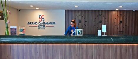 Mostrador de recepción en el Hotel Grand Cantagua, con un panel de pared de madera, un reloj y una pantalla digital.