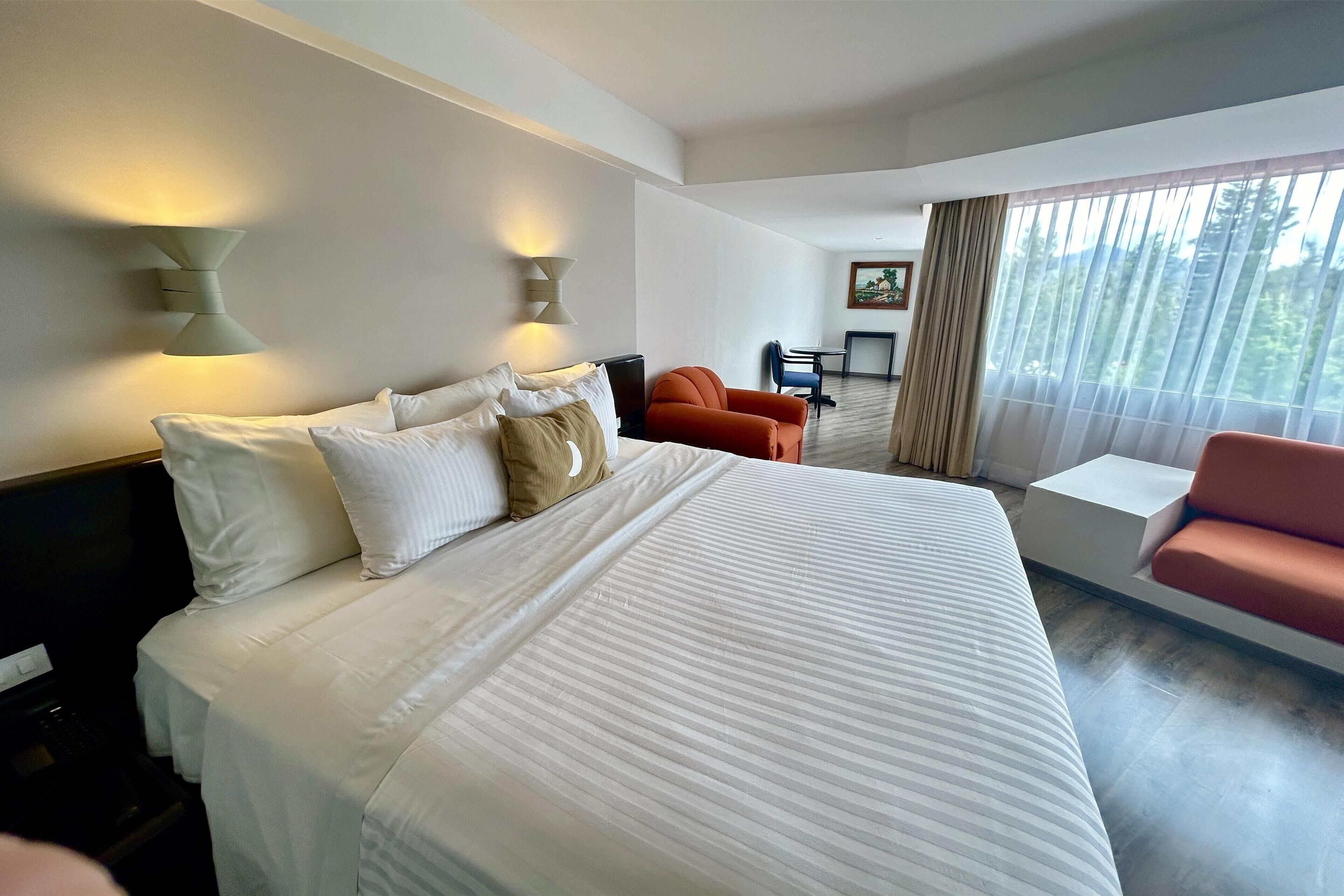 A hotel room with a large bed, two wall-mounted lights, a desk with a chair, and a view of the outdoors through sheer curtains.