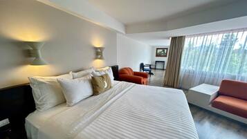 Quarto de hotel com uma cama grande, duas luzes fixadas na parede, uma escrivaninha com cadeira e vista para o exterior através de cortinas leves.