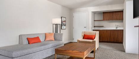 Una sala moderna con un sofá gris, una mesa de centro de madera y un sillón color beige. Hay una cocina con armarios de madera y una encimera blanca.