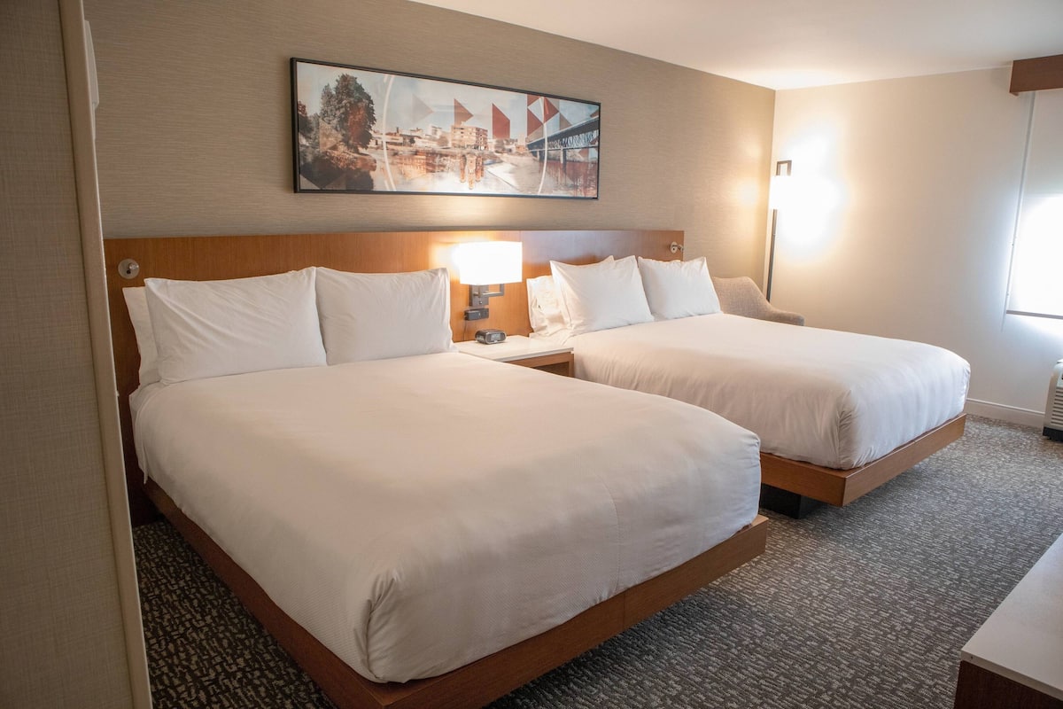 Habitación de hotel con dos camas, cabecera, lámpara, cuadro y suelo alfombrado.