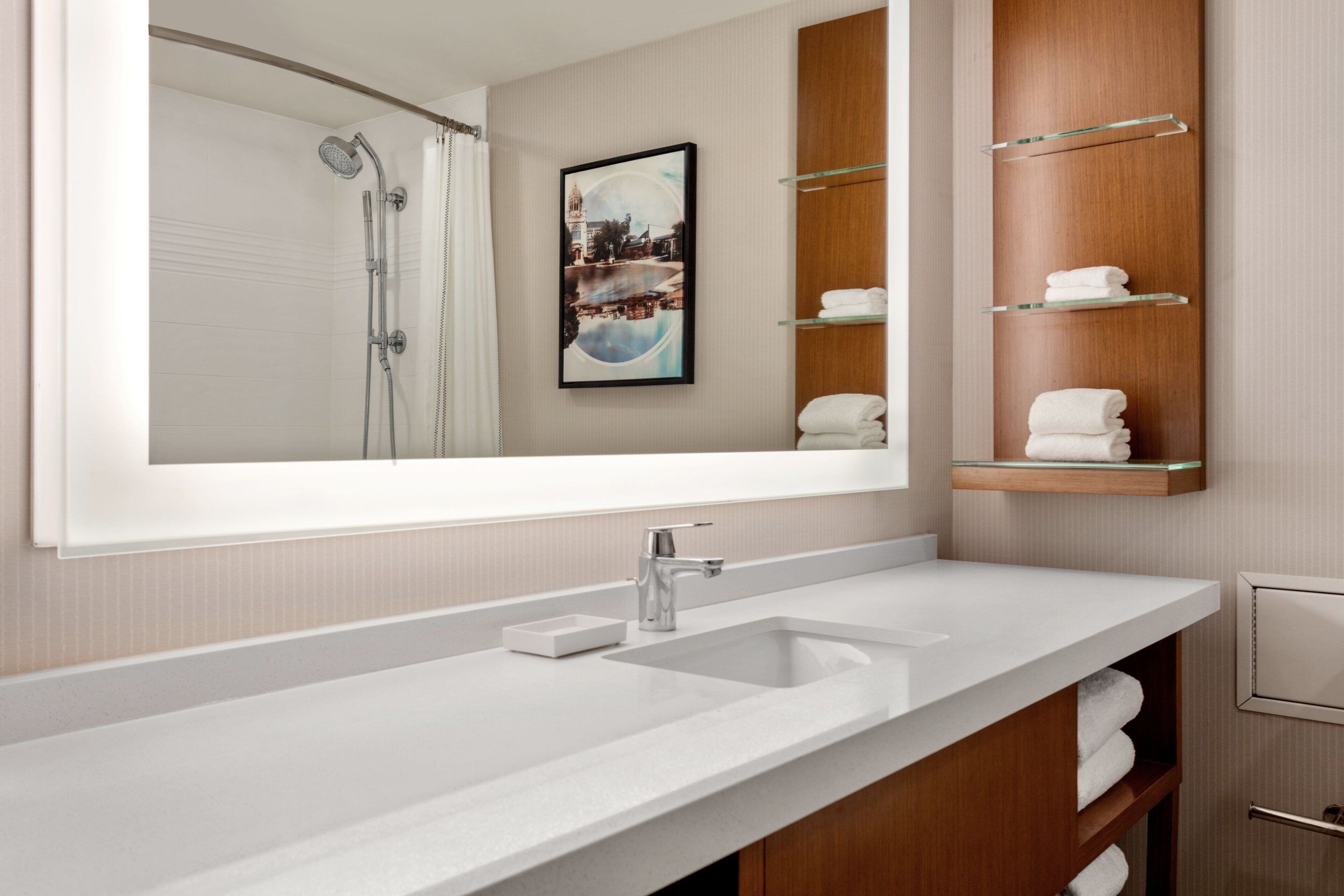 A modern bathroom with a large mirror, a white sink, a shower, and a wooden vanity with glass shelves holding towels.