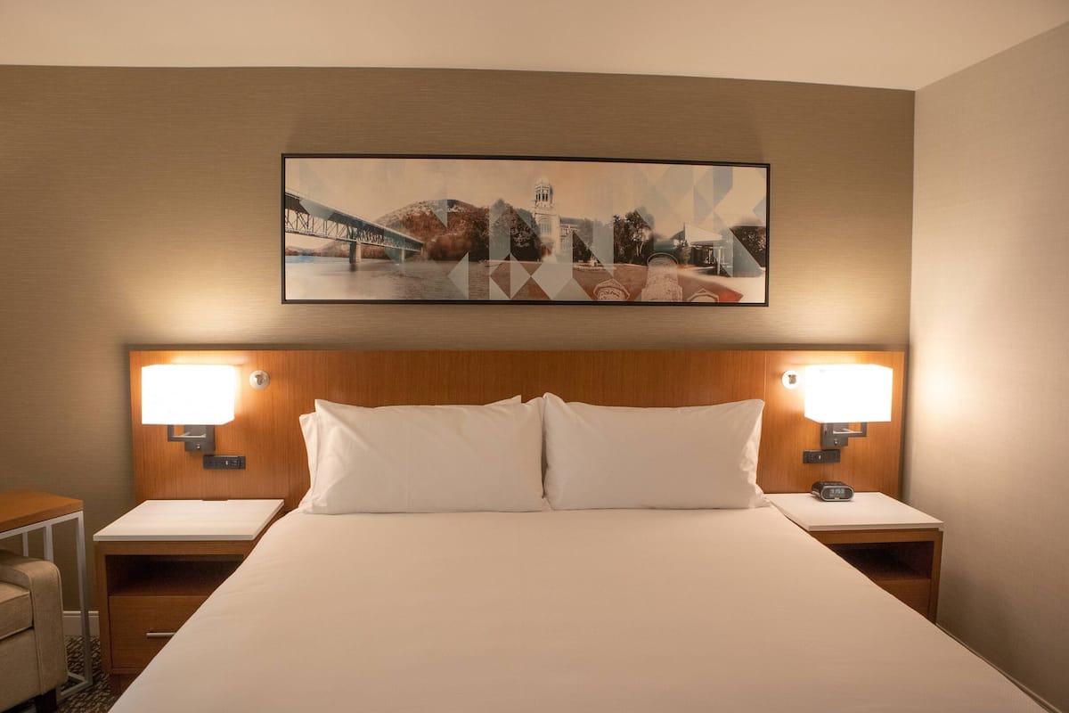 Una habitación de hotel con una cama grande, dos lámparas de noche, una mesita de noche y un cuadro con una vista urbana en la pared.