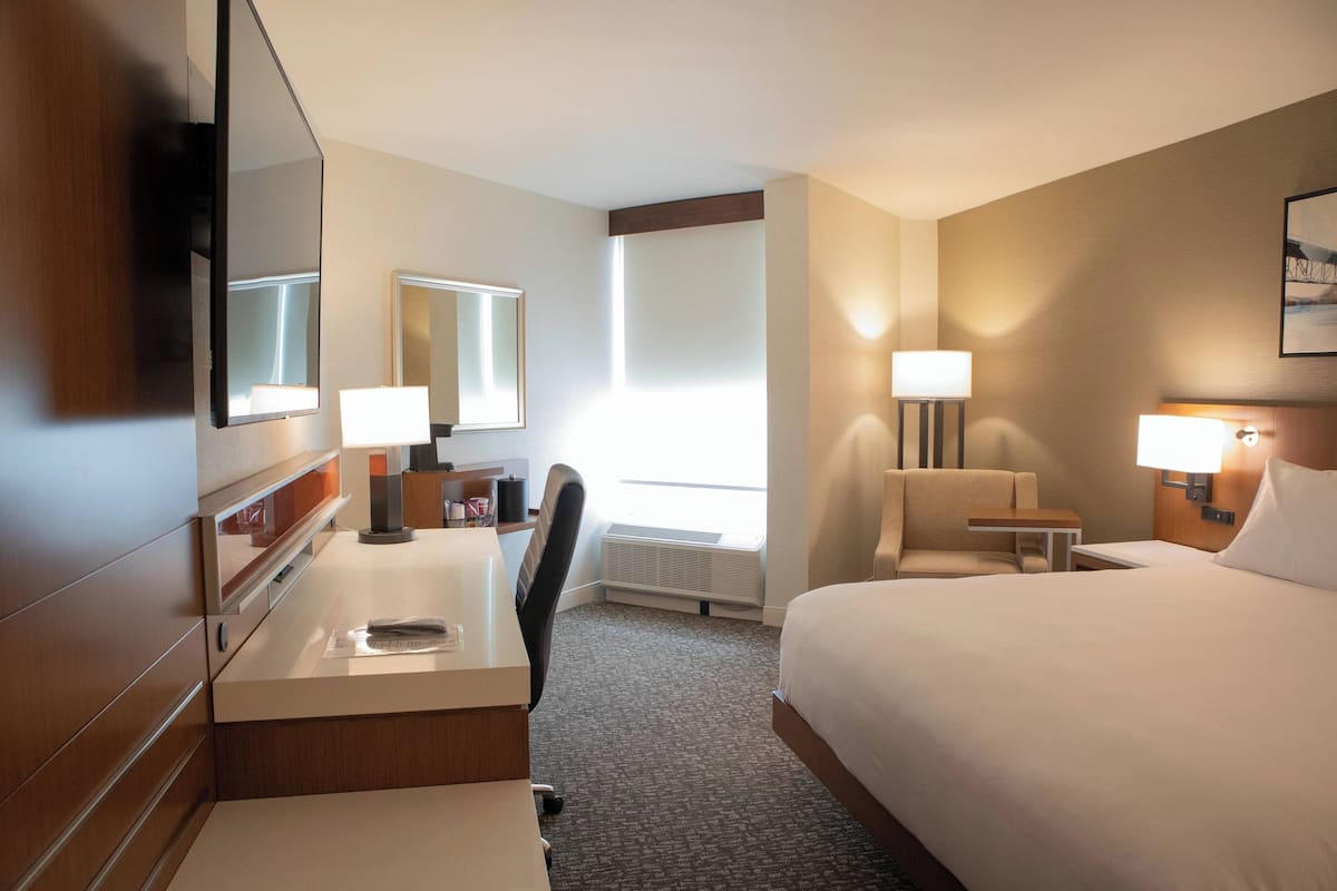 Habitación de hotel con cama, escritorio, silla, dos lámparas de noche, un espejo grande y una ventana.