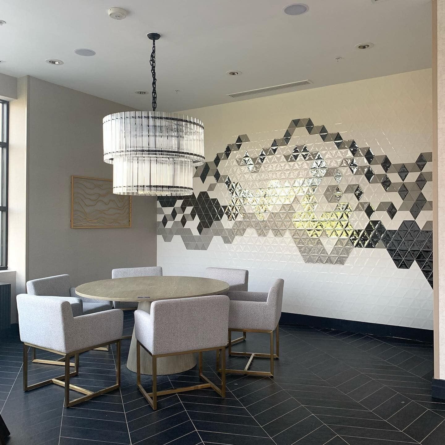 A modern dining area with a round table, six chairs, a chandelier, and a geometric wall design.