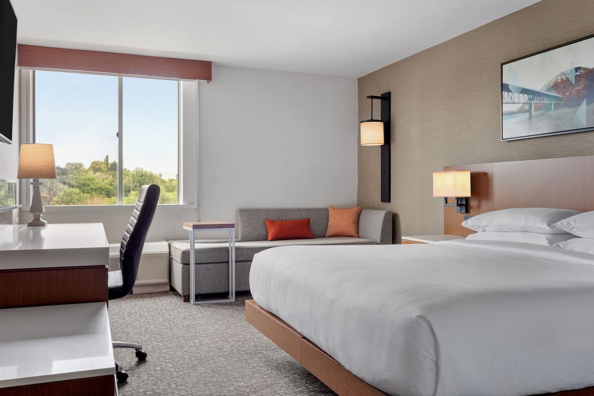 Habitación de hotel con una cama grande, un escritorio, una silla, un sofá y una ventana con vistas a zonas verdes.