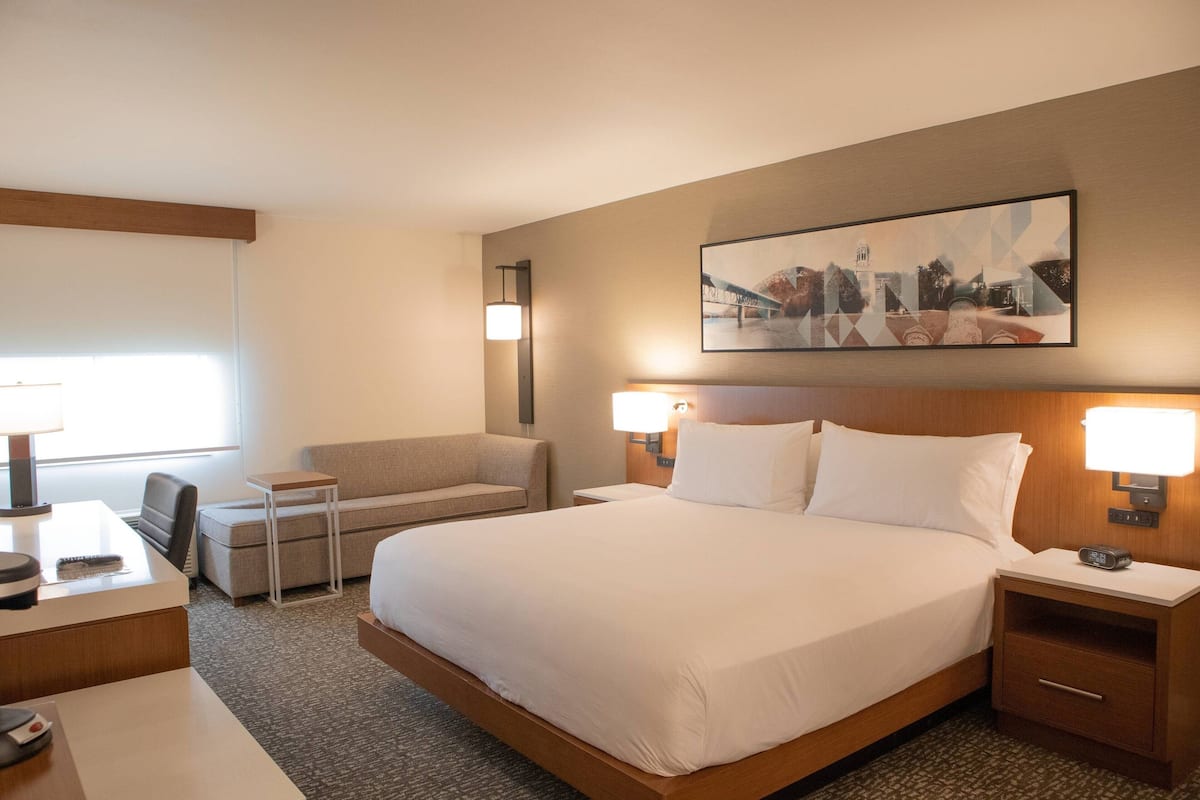 Habitación de hotel con una cama grande, dos mesitas de noche con lámparas, un escritorio con una silla, un pequeño sofá y un cuadro en la pared.