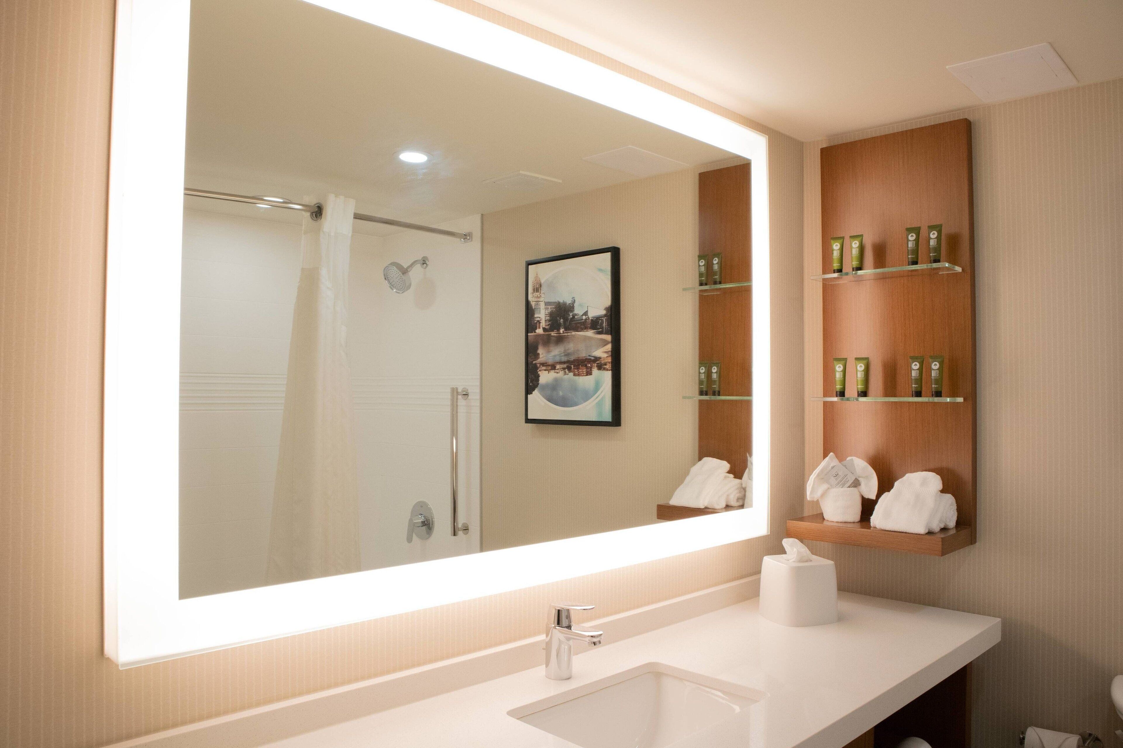 A modern bathroom with a large mirror, a sink, and a shower. There are toiletries on a shelf and a framed picture on the wall.