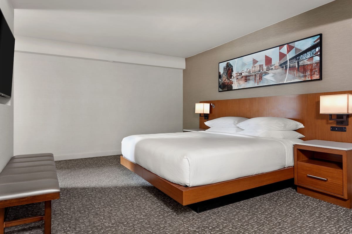 Una habitación de hotel con cama, mesitas de noche, un banco, un cuadro en la pared y un televisor.