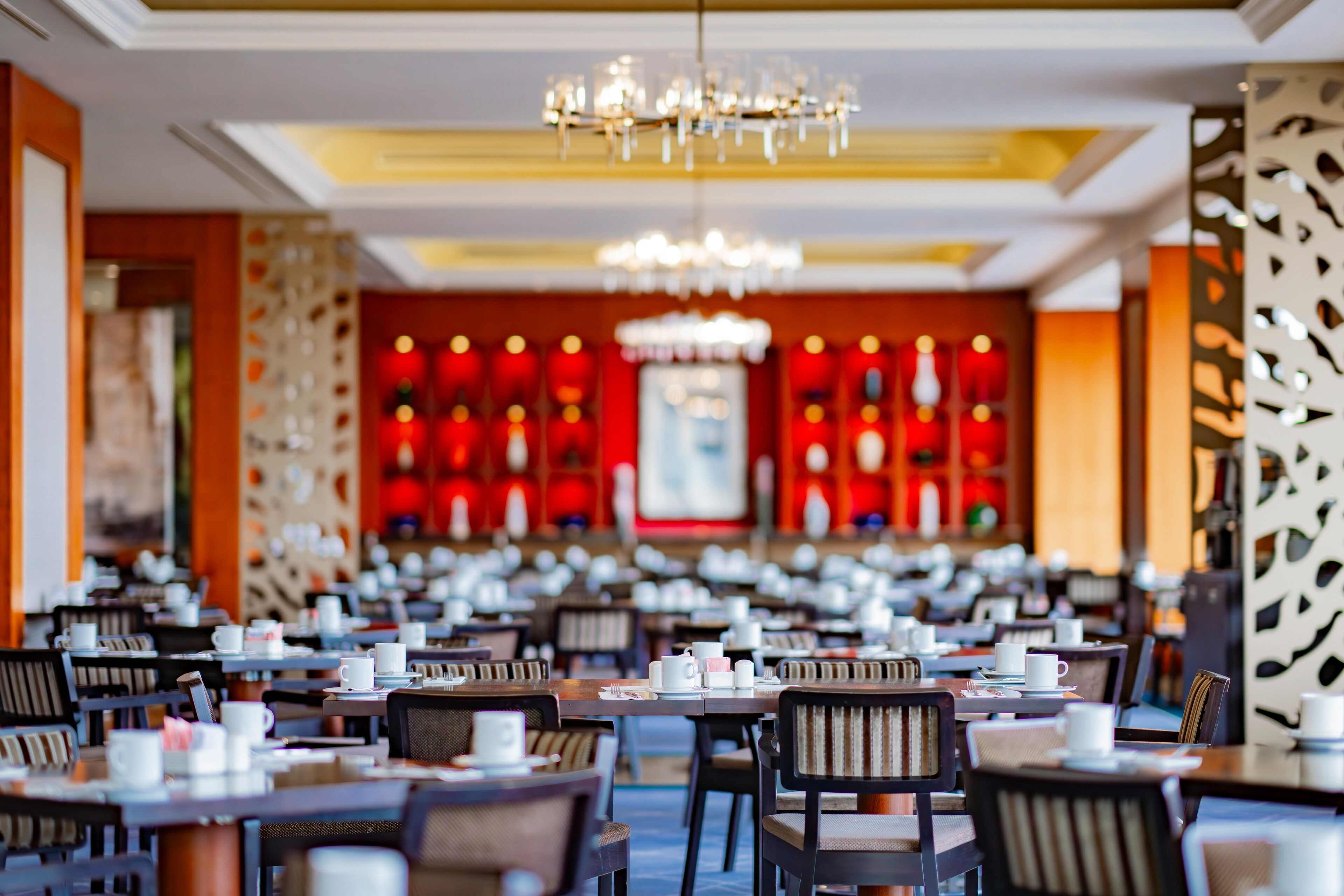 A spacious restaurant with multiple tables and chairs, a chandelier, and a red decorative wall in the background.