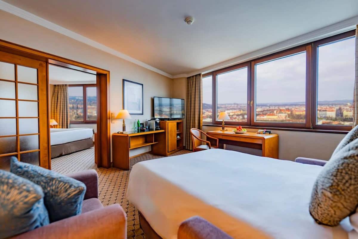 A hotel room with a bed, desk, chair, TV, and large windows with a city view.