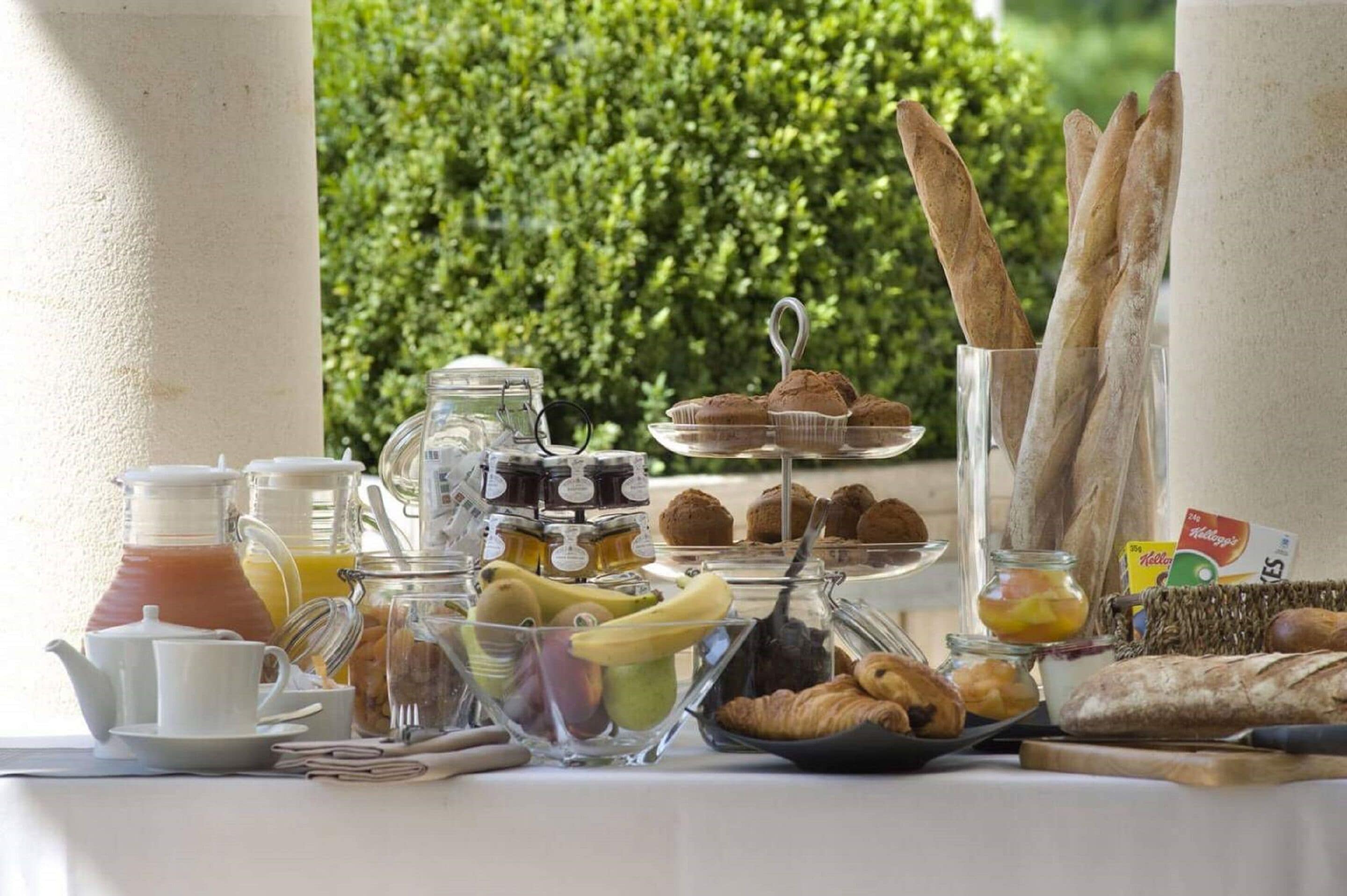 A breakfast buffet with various food items and beverages, including pastries, bread, and fruits, set against a backdrop of greenery.