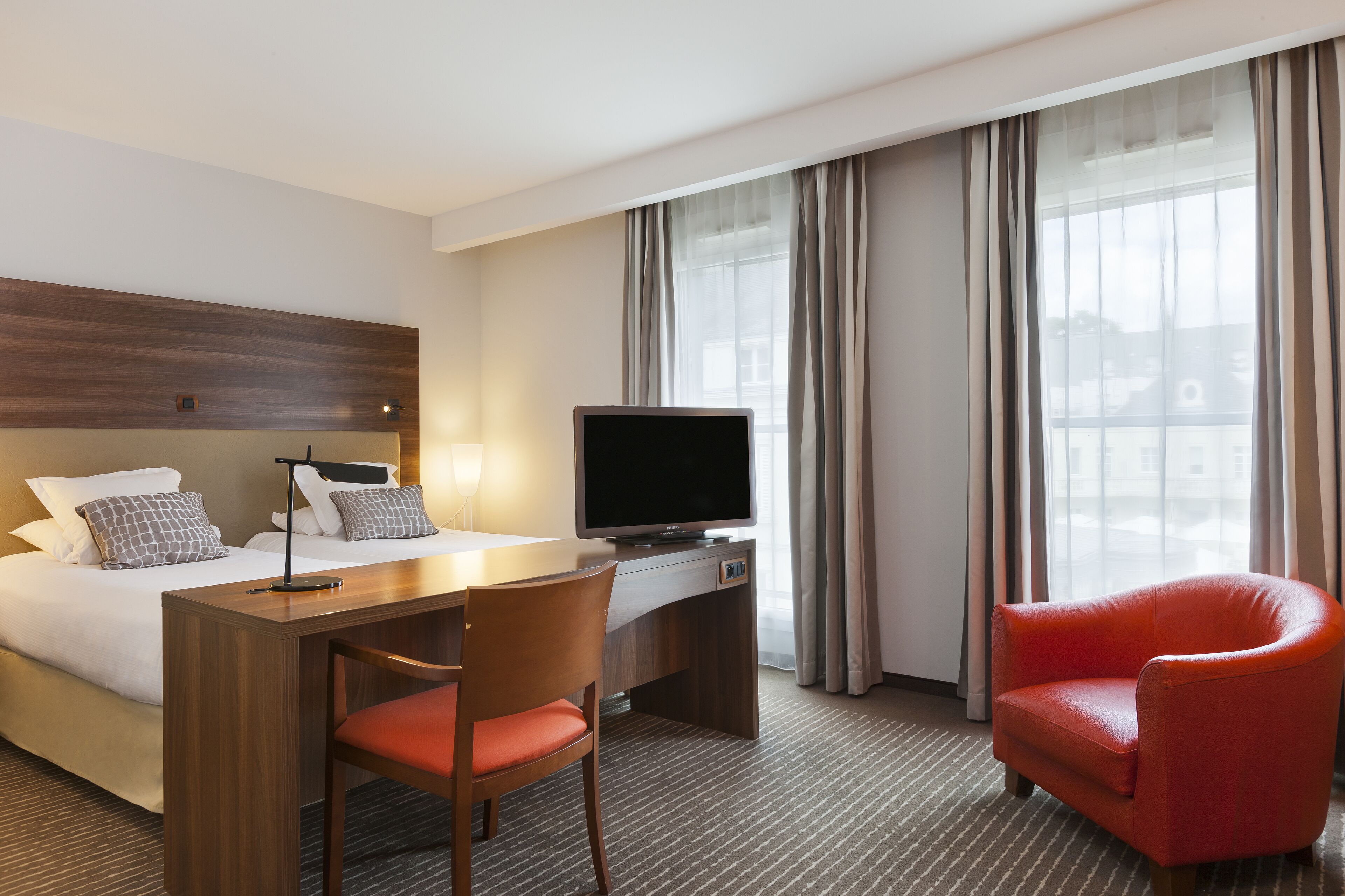 A hotel room with a bed, desk, TV, and red chair.