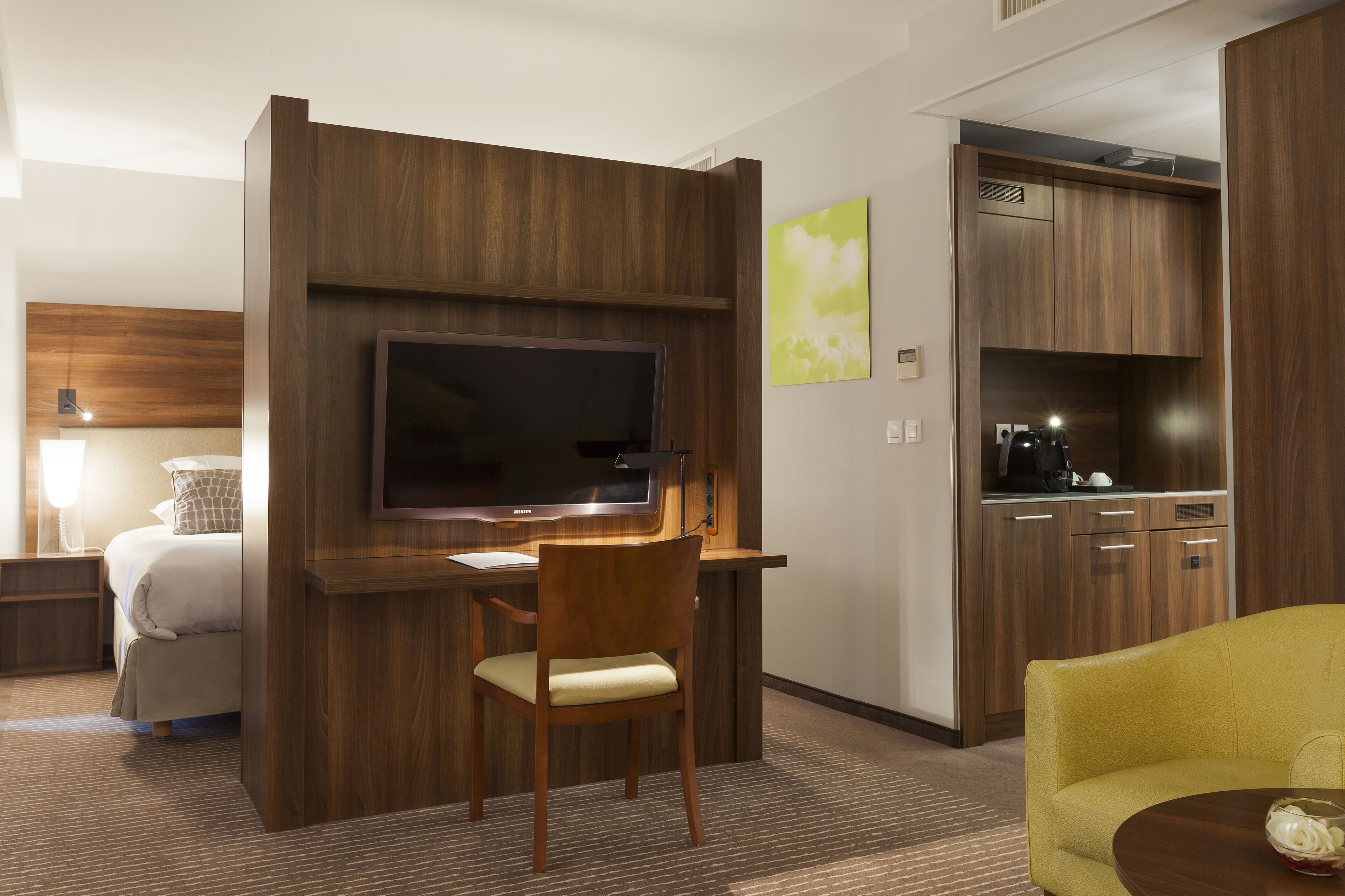 A hotel room with a bed, a desk with a chair, a television, a kitchenette, and a yellow armchair.
