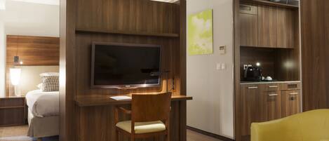 A hotel room with a bed, a desk with a chair, a television, a kitchenette, and a yellow armchair.