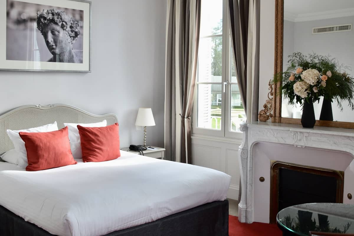 Une chambre d’hôtel avec un grand lit, une cheminée, un miroir et une fenêtre avec des rideaux.