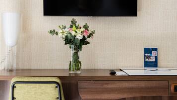 A hotel room with a wooden desk, a green chair, a flat-screen TV mounted on the wall, a vase with flowers, a white telephone, and a trash can.