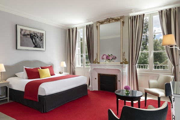 A hotel room with a large bed, a fireplace, a sitting area with a table and chairs, and a large window with curtains.