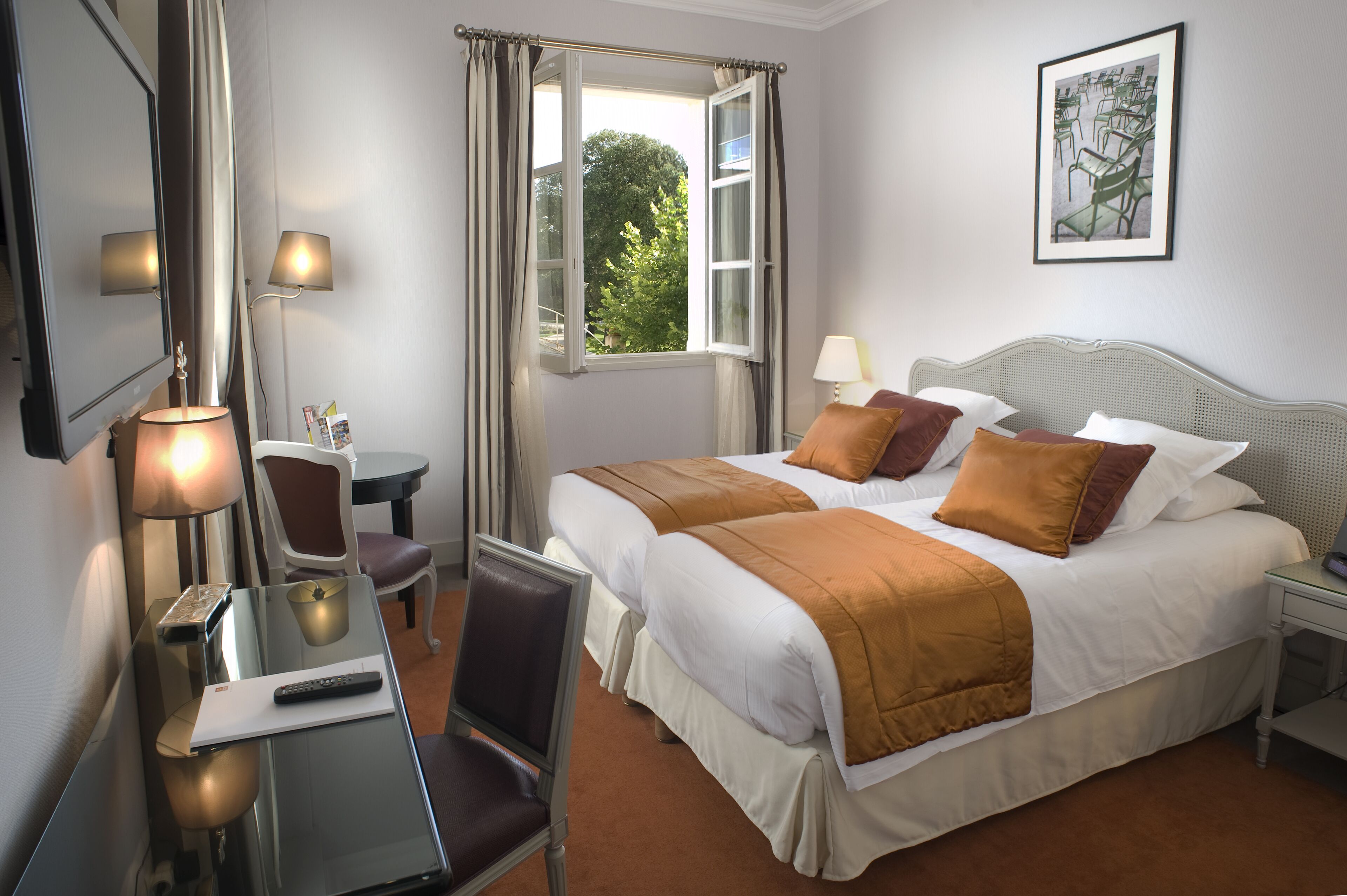 A hotel room with a bed, bedside tables, a desk with a chair, a lamp, and a window with curtains.