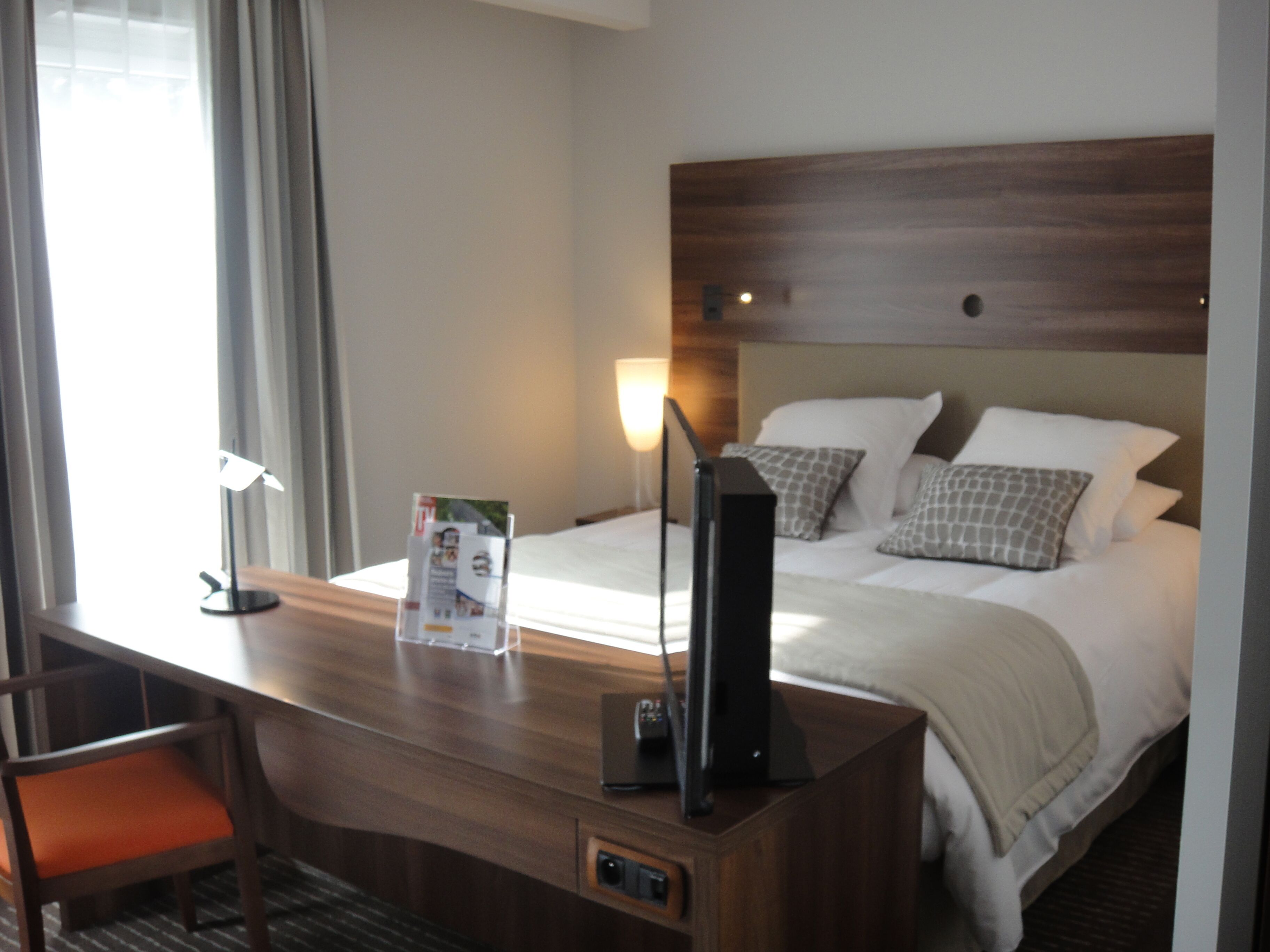 A hotel room with a wooden desk, a chair, a bed with patterned pillows, and a nightstand with a lamp and a remote control.