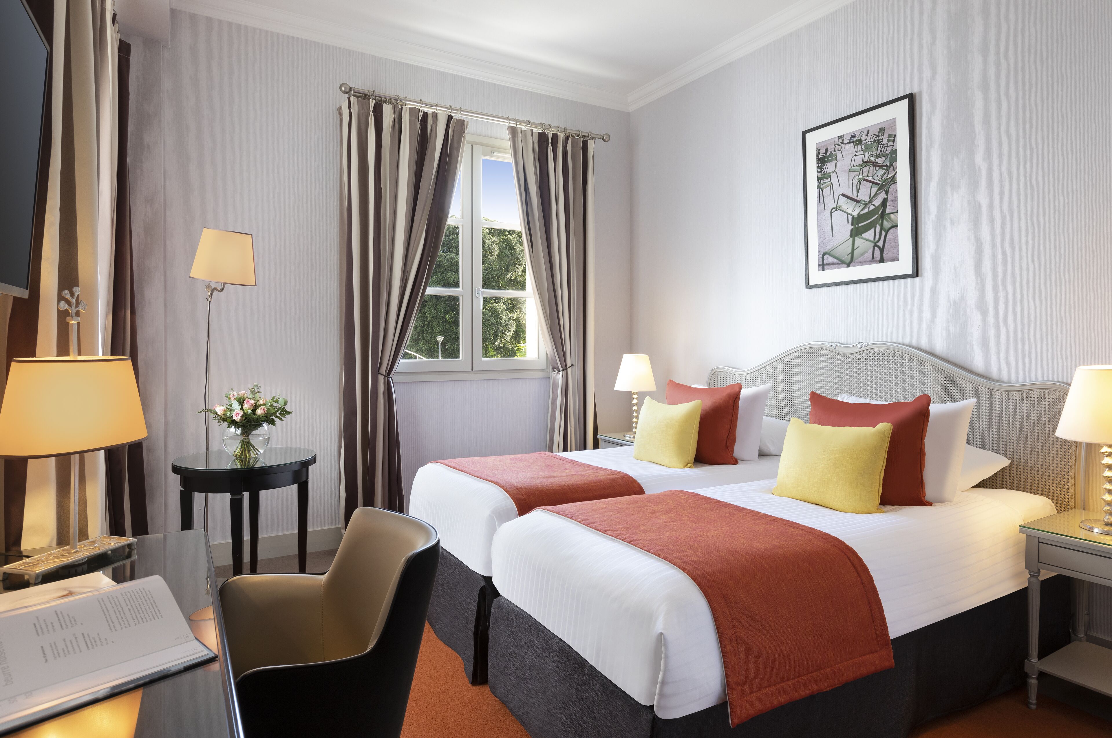Une chambre d’hôtel avec deux lits, un bureau avec une chaise, une petite table avec un vase de fleurs et une fenêtre avec des rideaux.