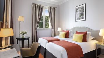 Habitación de hotel con dos camas, un escritorio con silla, una mesita con un jarrón de flores y una ventana con cortinas.