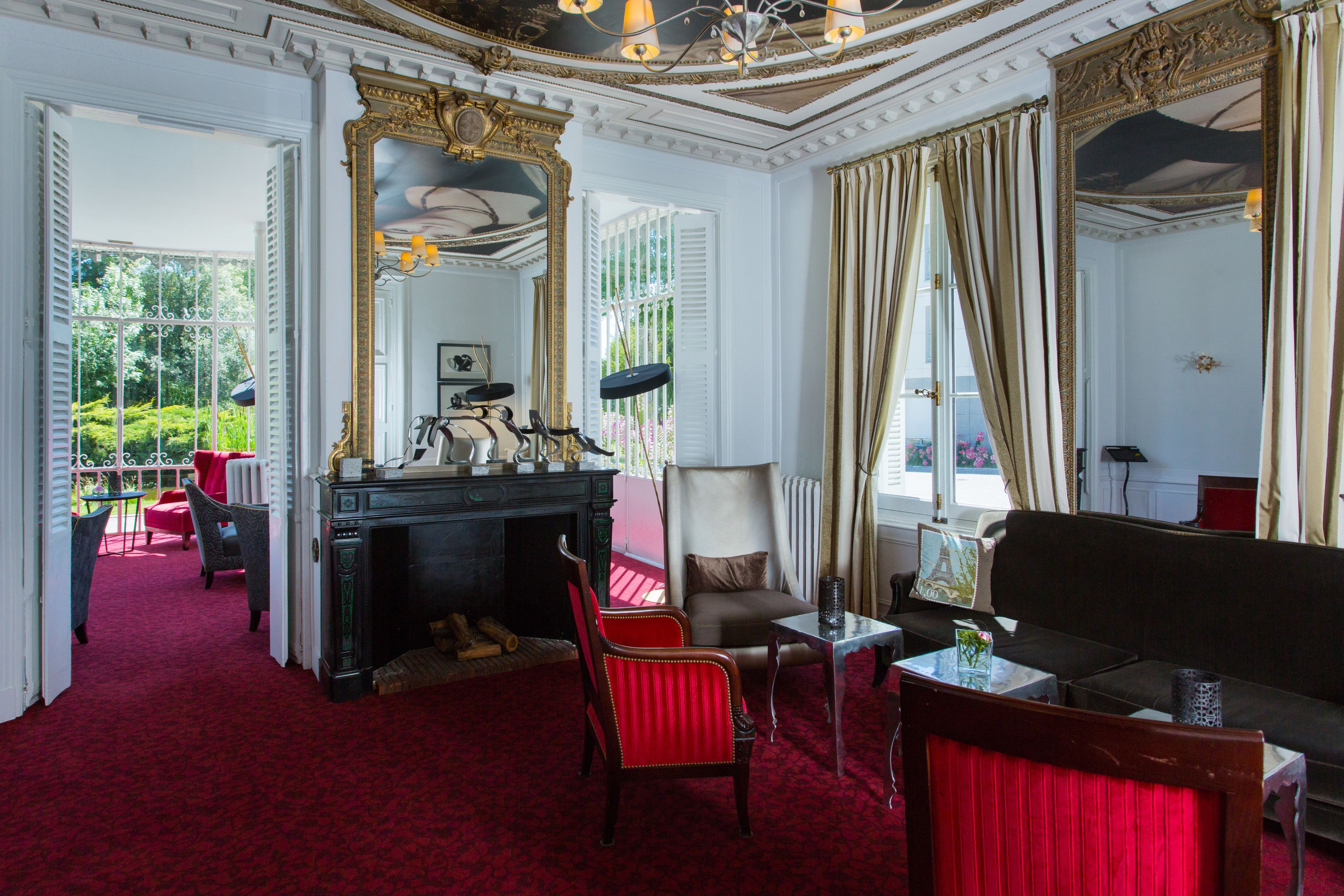 Une pièce avec une cheminée, un canapé et un fauteuil. Il y a une fenêtre avec des rideaux, et de là, on peut admirer la verdure à l’extérieur.