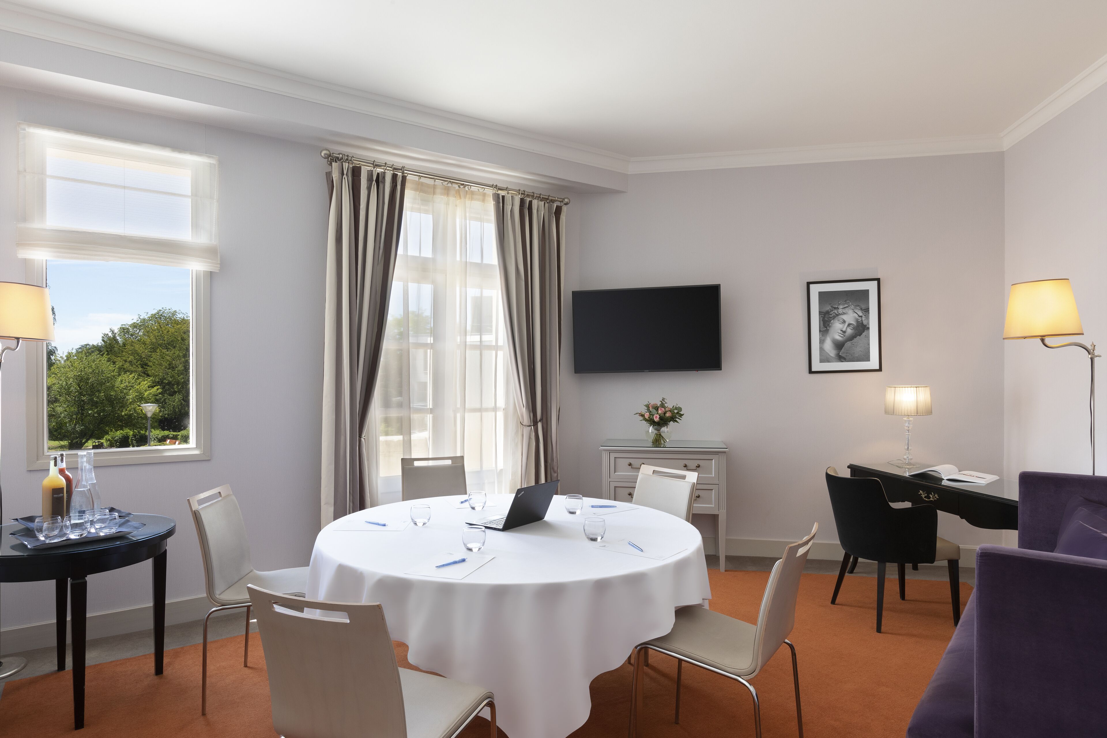 Habitación de hotel con una mesa de comedor redonda, sillas, un televisor de pantalla plana, un escritorio con lámpara y una ventana con cortinas.