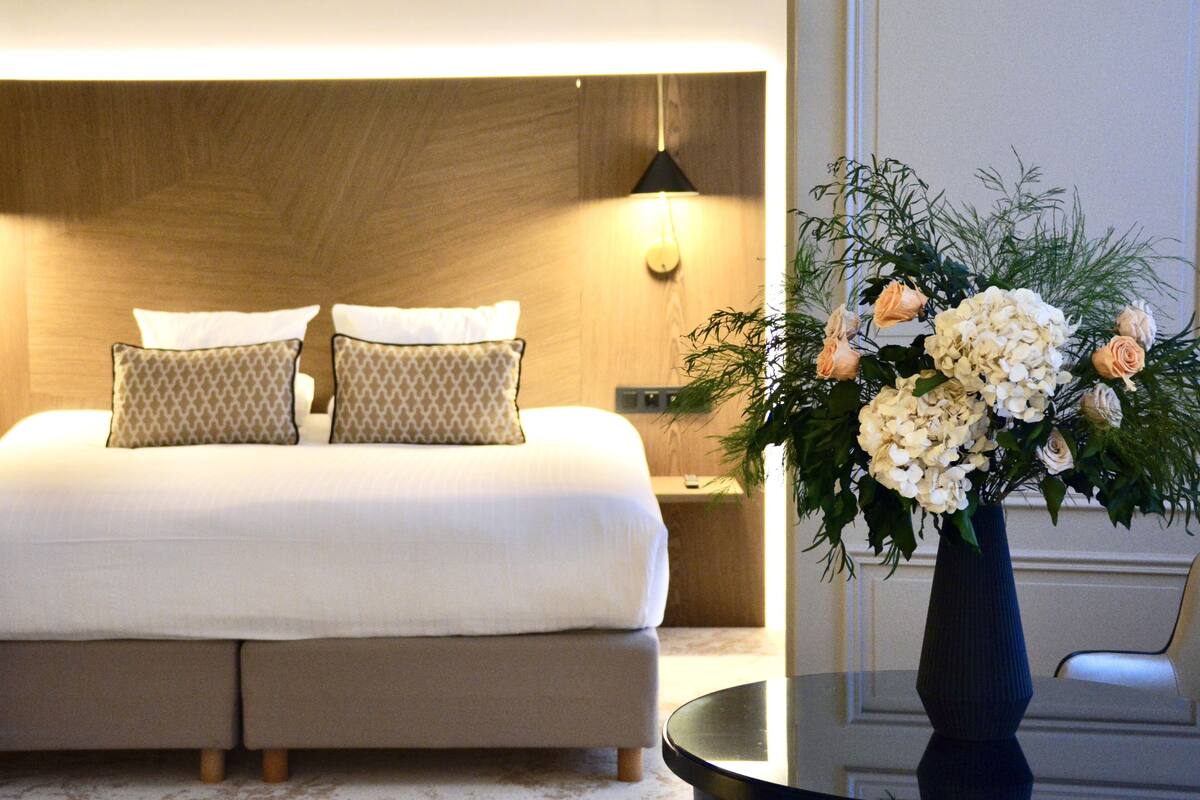 Un lit bien fait, agrémenté d’oreillers à motifs, une lampe de chevet et un vase de fleurs posé sur une petite table à côté du lit.