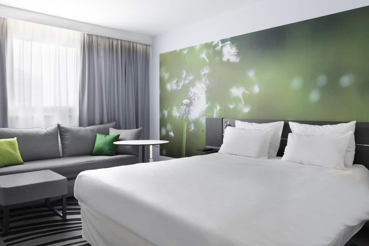 Une chambre dâhĂŽtel moderne, dotĂ©e dâun grand lit, dâun canapĂ©, dâune petite table et dâun mur vert sur lequel est projetĂ©e lâimage dâun pissenlit.