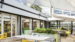 An outdoor seating area with tables and chairs, a wooden deck, and a glass enclosure with plants. - Novotel Paris Charles de Gaulle Airport (Tremblay-en-France)