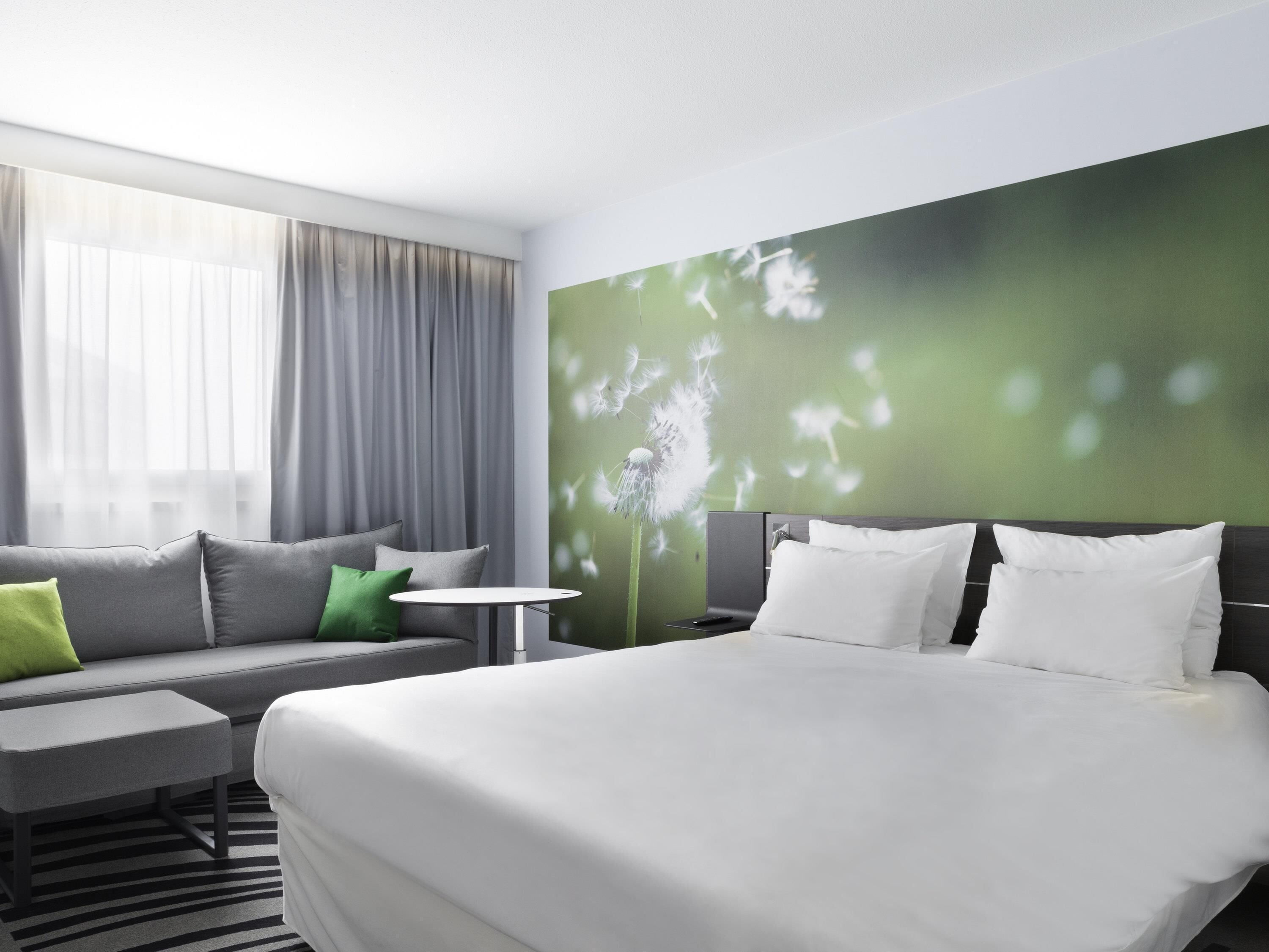 Ein modernes Hotelzimmer mit einem großen Bett, einer Couch, einem kleinen Tisch und einer grünen Wand mit einem Löwenzahnbild.