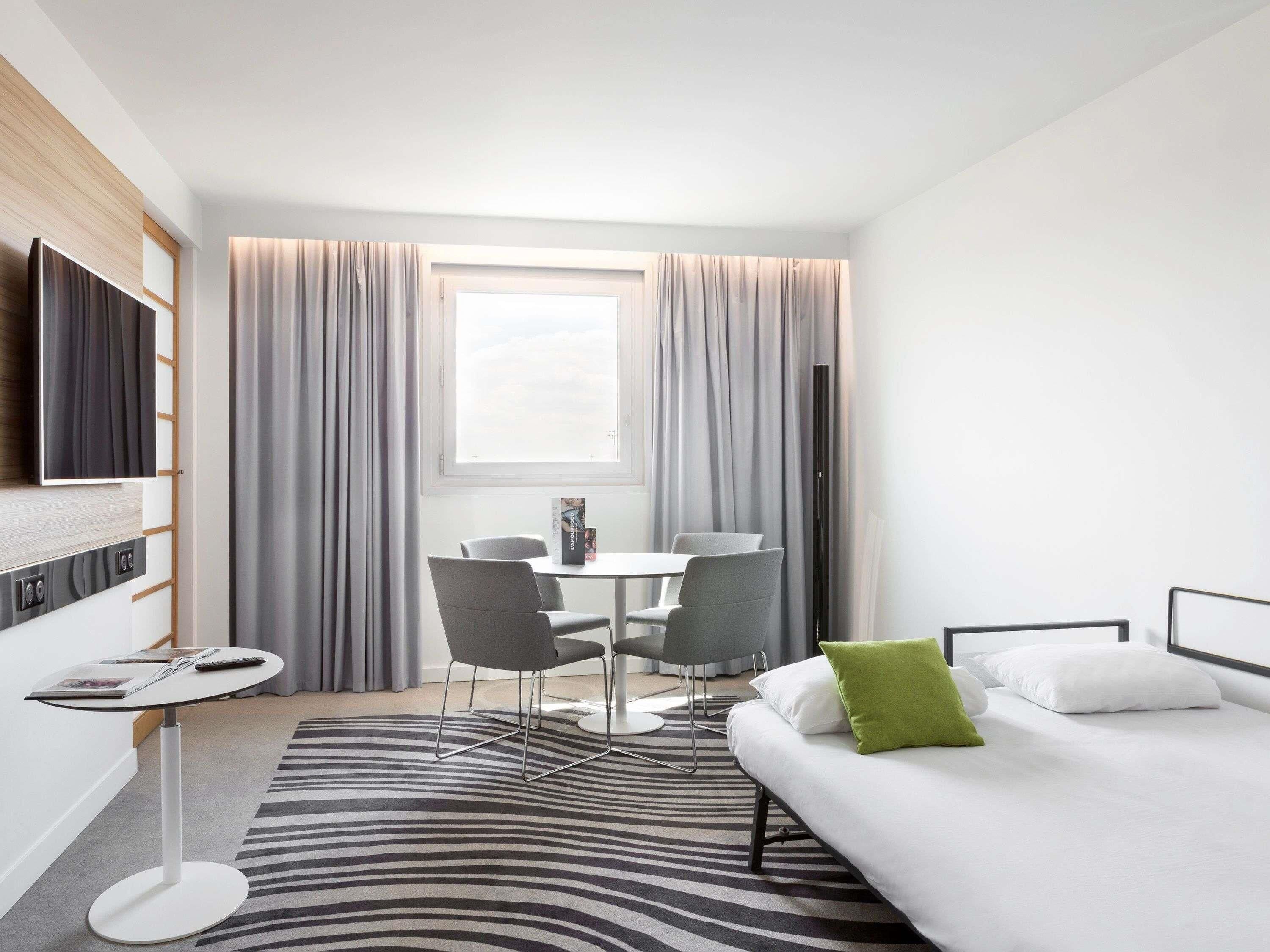 A modern hotel room with a bed, a desk with two chairs, a flat-screen TV, and a window with curtains.