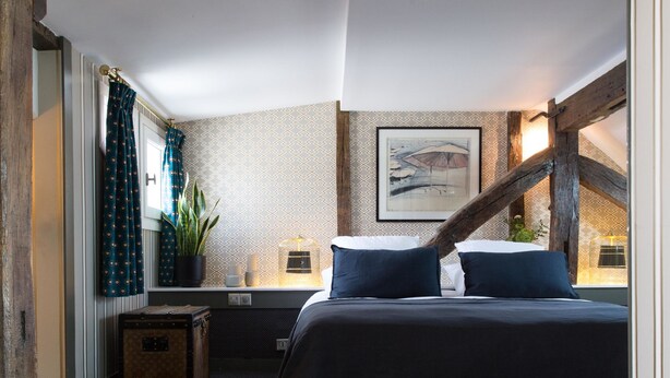A bedroom with a bed, a window with curtains, a plant, a bedside table, and a painting on the wall. - Relais Saint Germain (Paris)