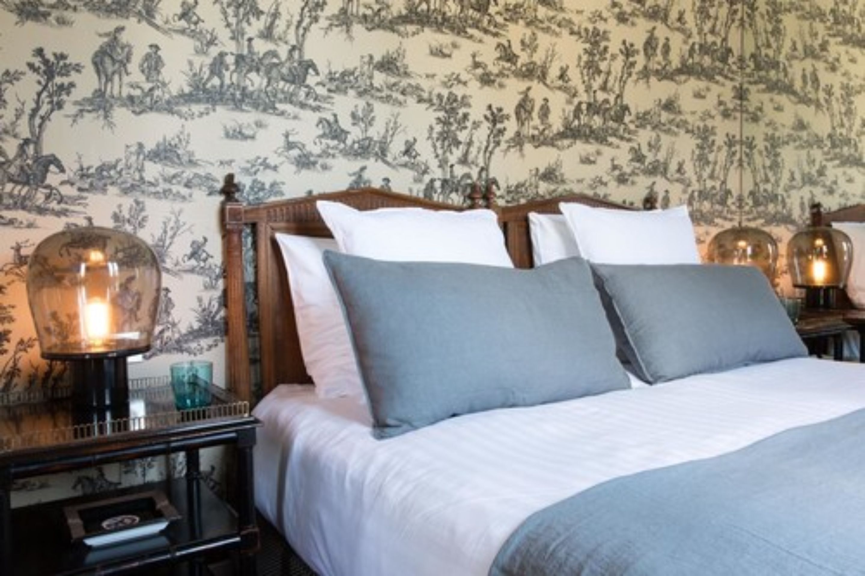 Una cama bien hecha con ropa de cama blanca y gris, un cabecero de madera y dos lámparas de noche con pantallas de vidrio transparente.