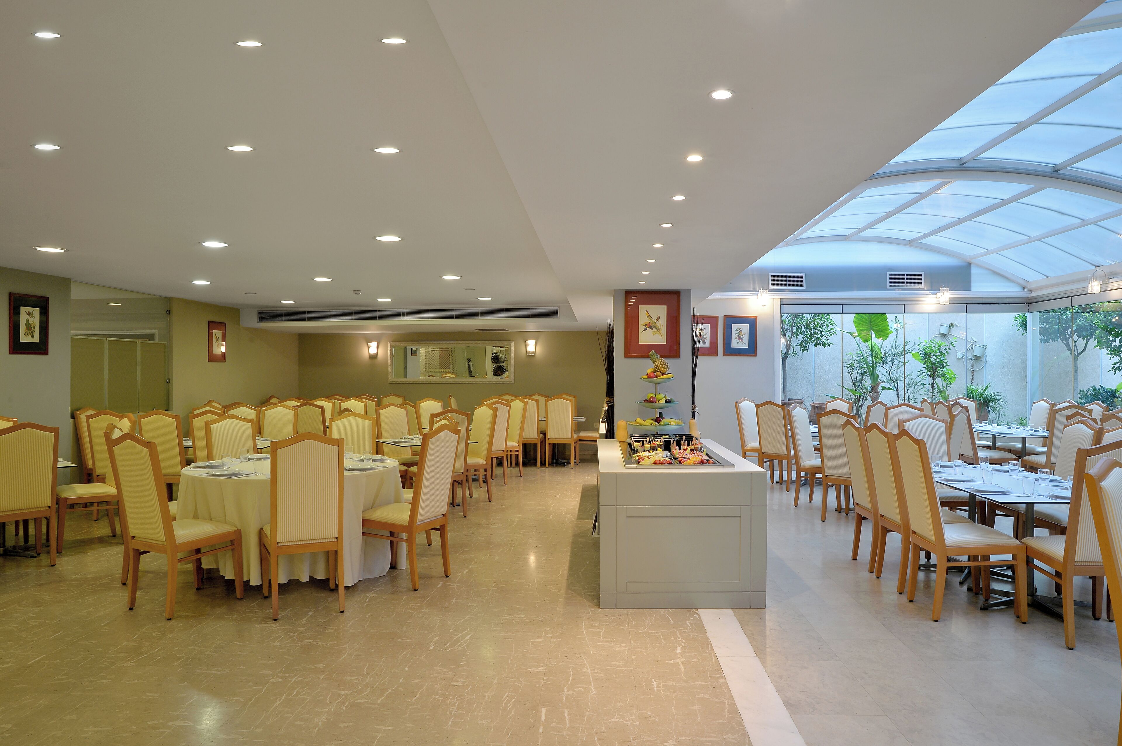 A spacious dining area with multiple tables and chairs, a central buffet station, and a large glass ceiling allowing natural light.