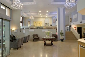 A spacious lobby with a reception desk, comfortable seating, a chandelier, and a staircase leading upstairs.