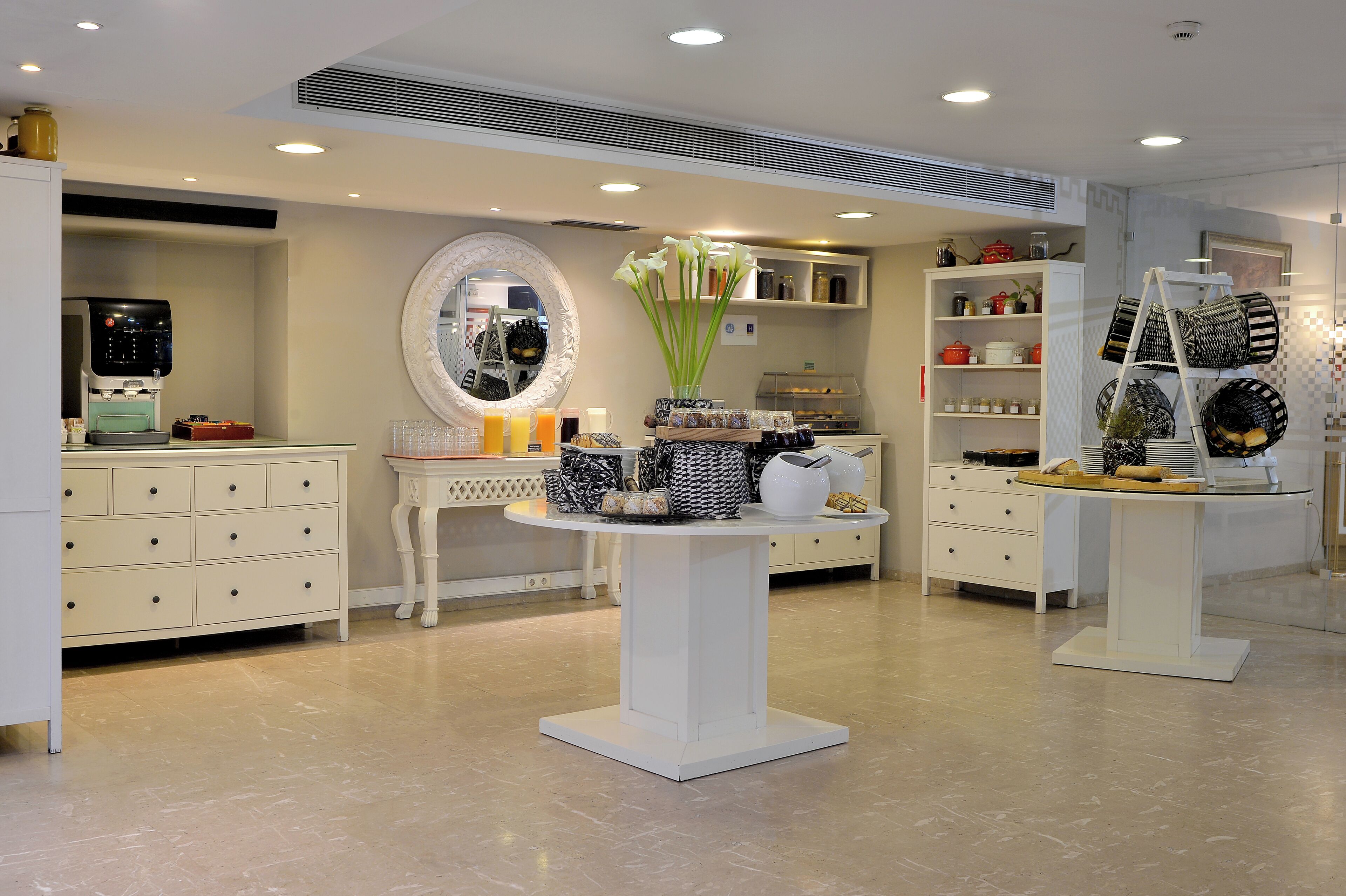 A modern hotel lobby with a coffee machine, a round mirror, a table with decorative items, and a display of bags.