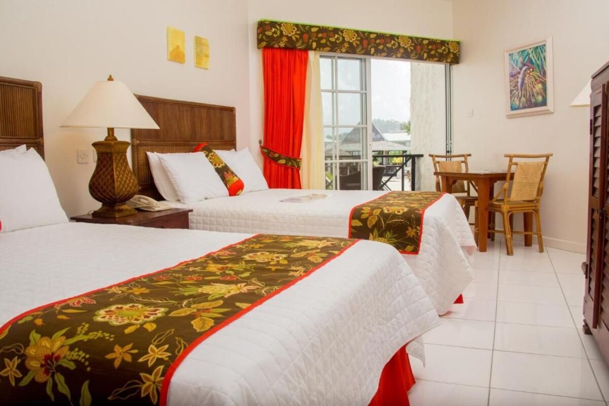 A hotel room with two beds, a dining table, chairs, a lamp, and a view of the outdoors.