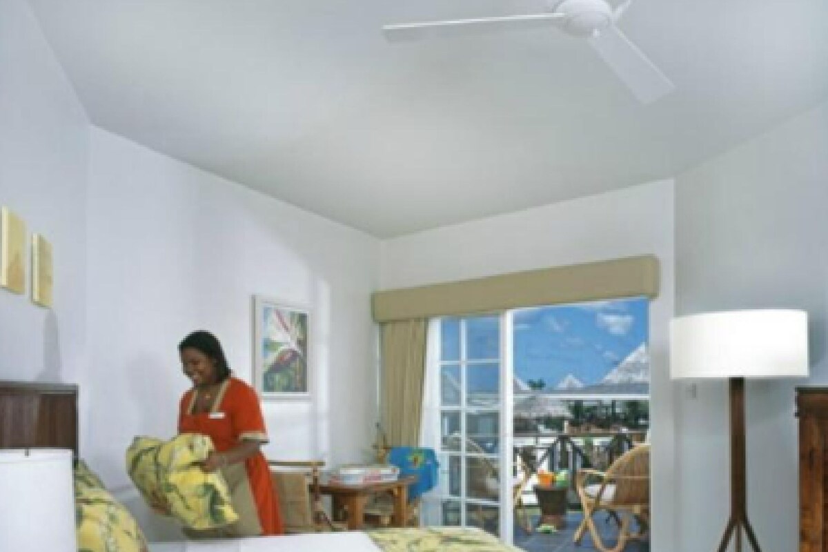A hotel room with a bed, a ceiling fan, a window with a view of the outdoors, and a person folding a blanket.