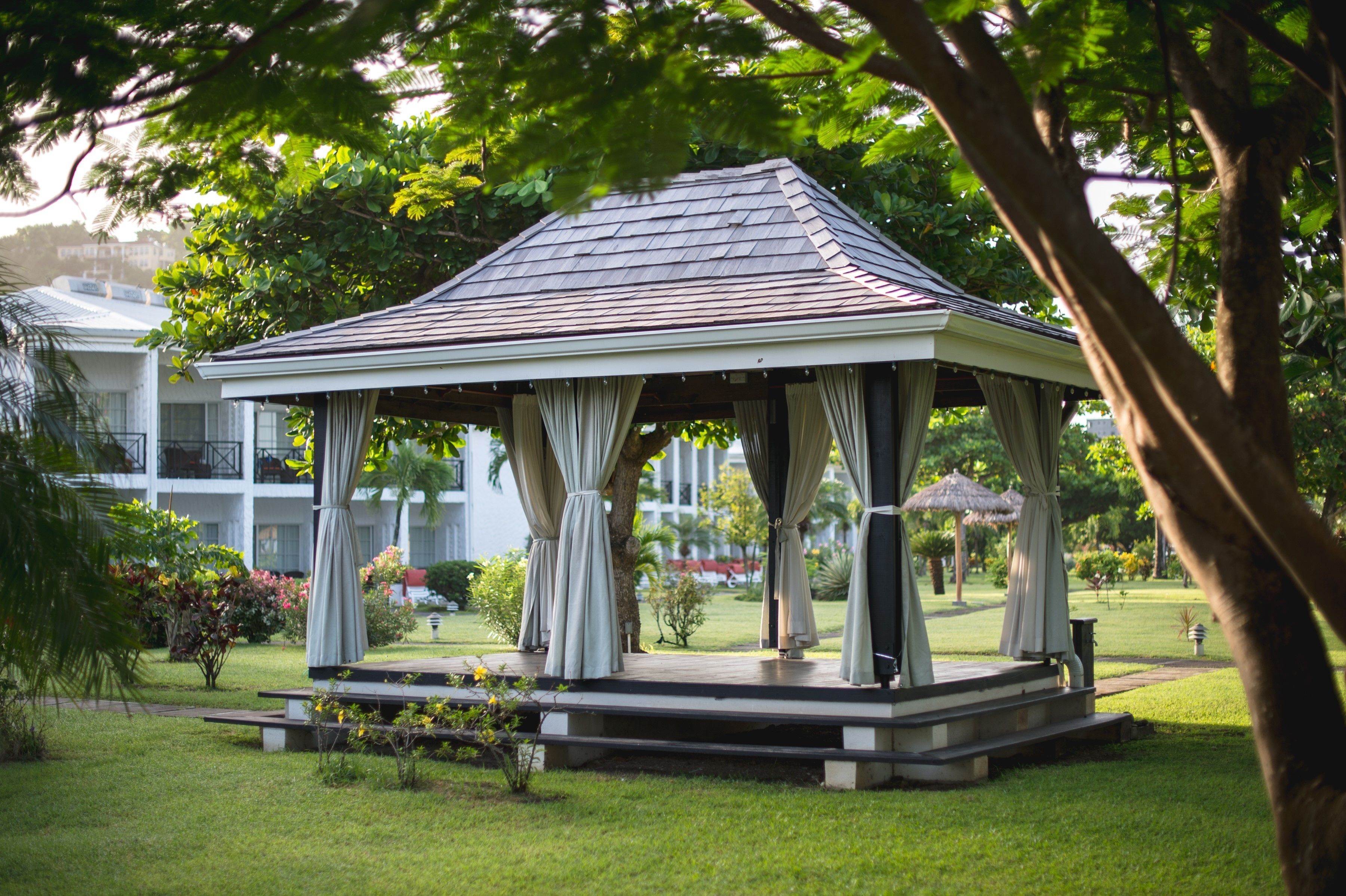 A gazebo with a tiled roof and white walls, surrounded by lush greenery and a well-maintained lawn.